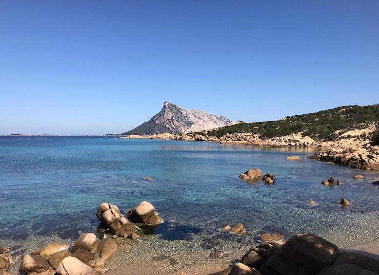 A clear coastal scene with a rocky shoreline, calm sea, and a prominent mountain in the background. 1