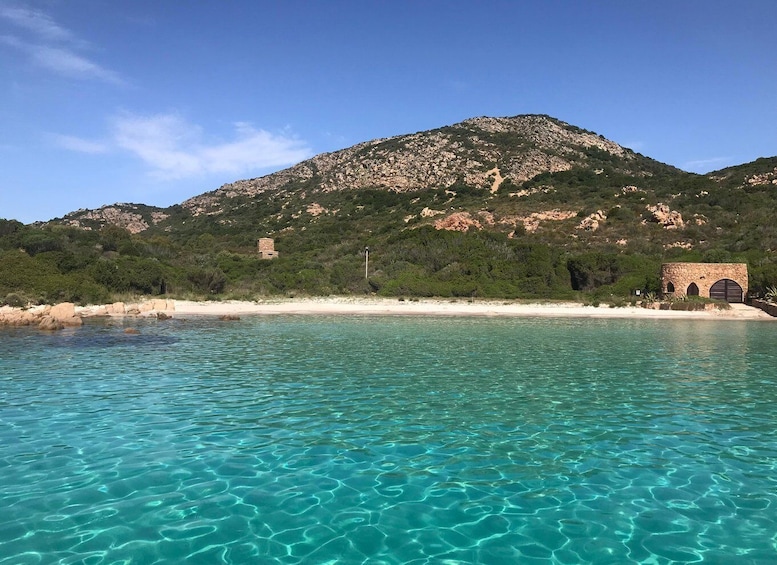 A coastal landscape with clear turquoise waters, a sandy beach, and a rocky hill with a small stone structure. 3