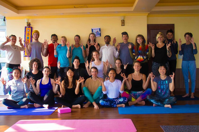 A group of people in a yoga class, sitting and standing on mats, with some in colorful attire. 5