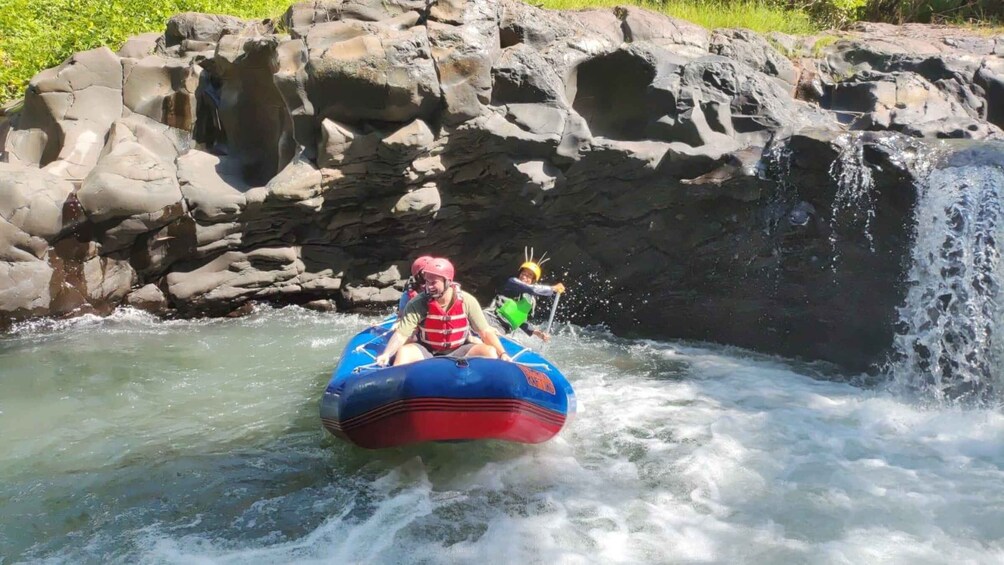 Two people in a blue inflatable raft navigating a river with a waterfall in the background. 3