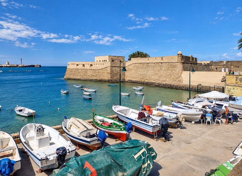 A coastal harbor with boats docked, a fortified wall, and a lighthouse in the background. 5