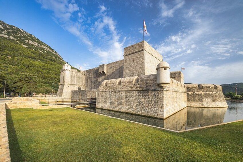 A historic stone fortress with a flag, surrounded by a moat and grassy area. 3