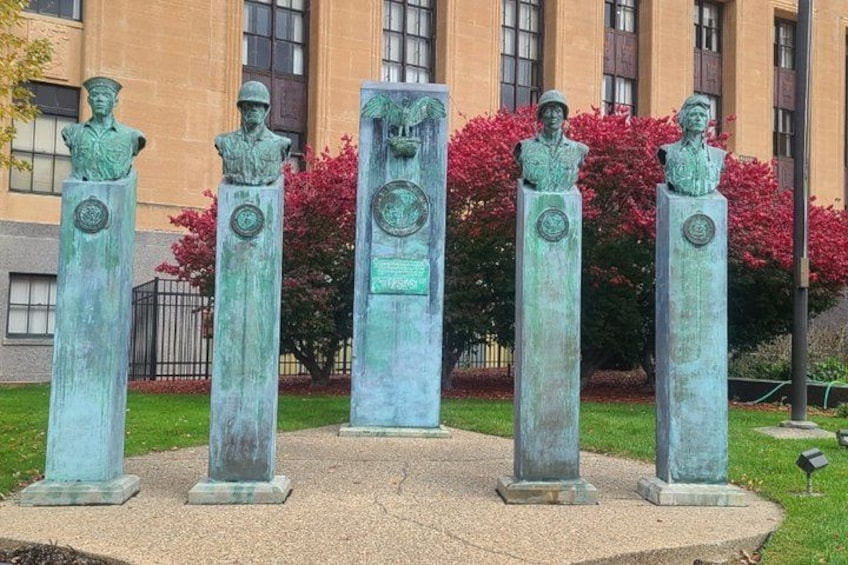 A series of bronze statues on pedestals in a landscaped area with a building in the background. 2