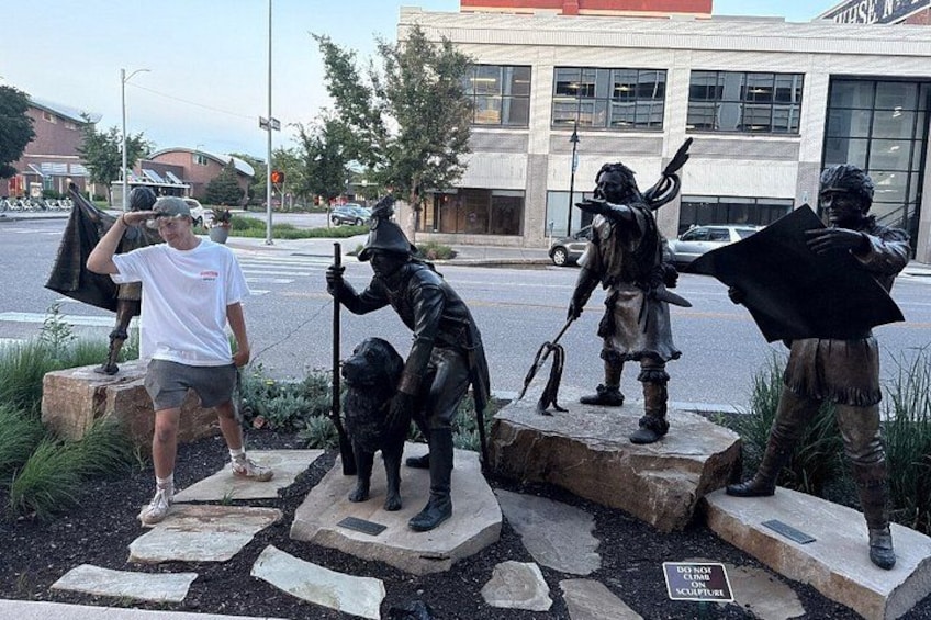 A statue of a man in a white shirt and shorts standing next to a statue of a Native American figure. 1
