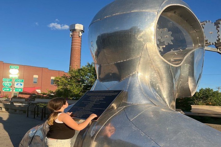 A large metallic sculpture with gears, a woman reading a plaque, and a red brick building in the background. 3