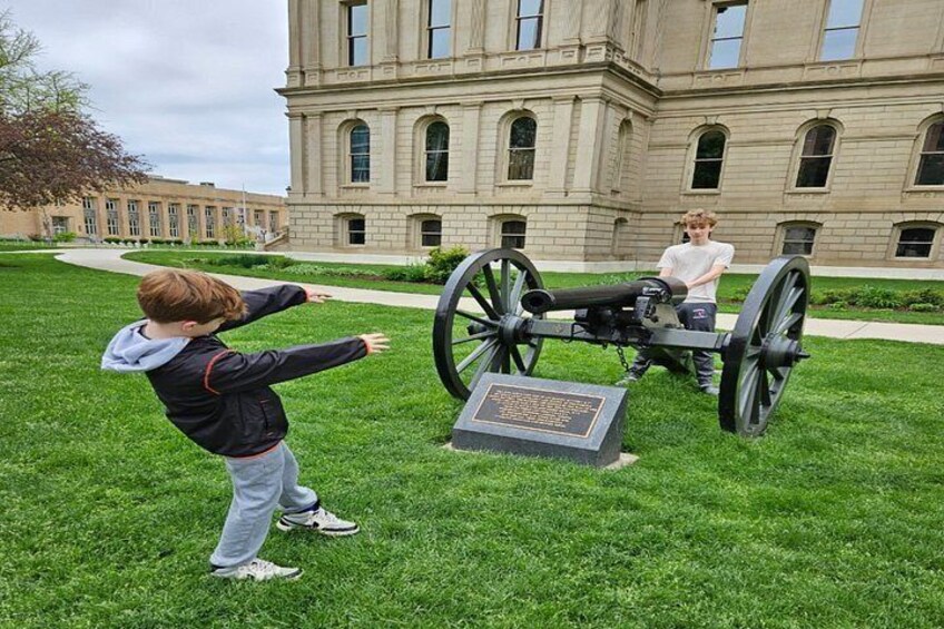 A child aiming a toy cannon at a historical cannon in a grassy area with a building in the background. 4