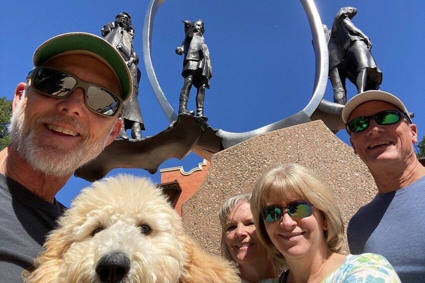 A group of people with a dog in front of a sculpture with figures on top. 3