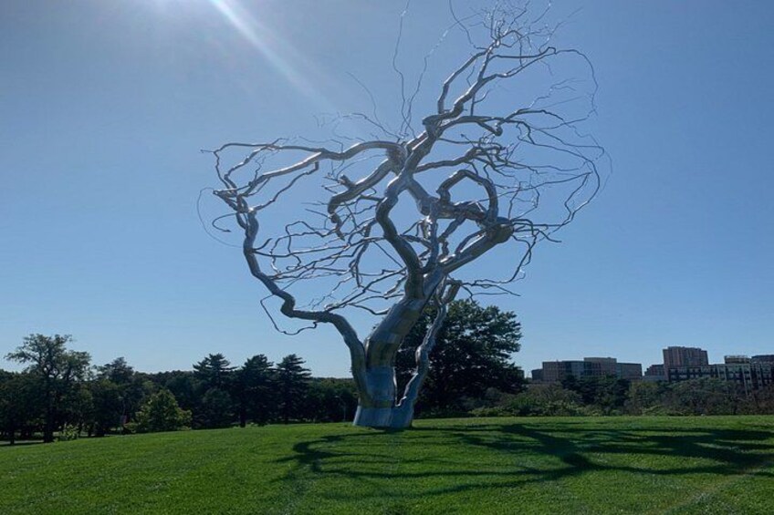 Una gran escultura metálica de un árbol en un campo cubierto de césped, con edificios al fondo. 2