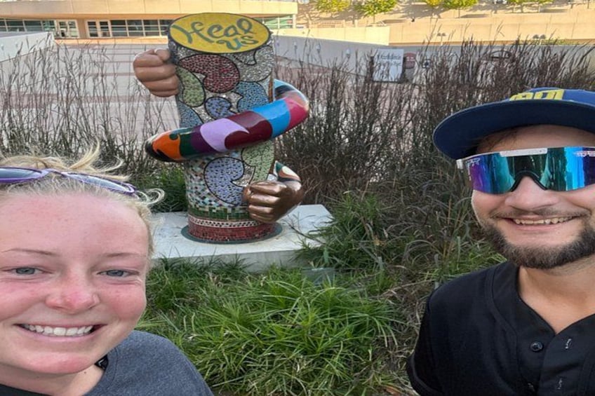 A sculpture of a hand holding a colorful object with the text 'Keep us' on it, set in a grassy area with a building in the background. 3