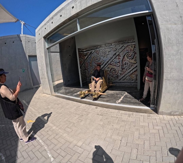 A person sitting on a wooden chair in front of a building with a patterned wall. 4