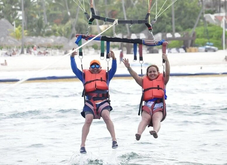 Two people parasailing over water, wearing life jackets and holding onto the parasail structure. 3