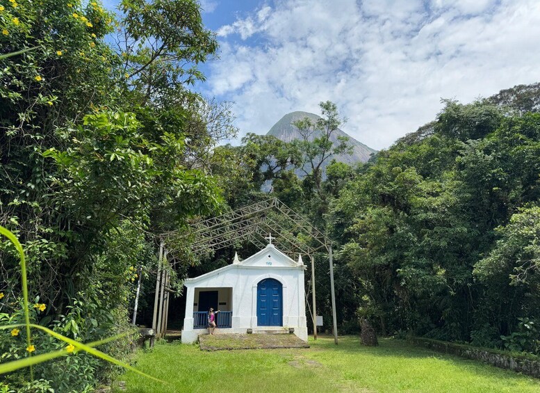 A white gazebo with a blue door in a lush green garden, surrounded by trees and a mountain in the background. 3