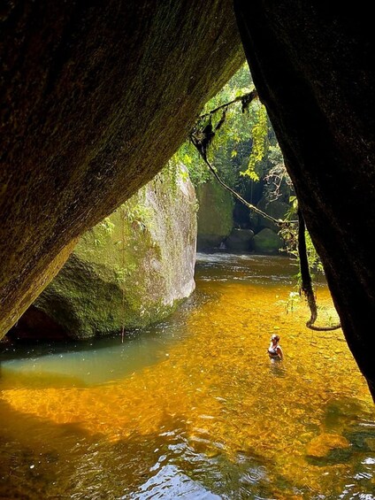 A person wading in a clear, shallow stream under a large, moss-covered rock formation. 4