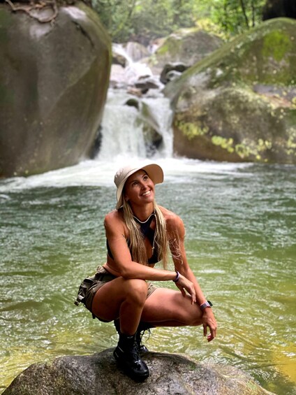 A person squatting on a rock by a flowing river with a waterfall in the background. 1