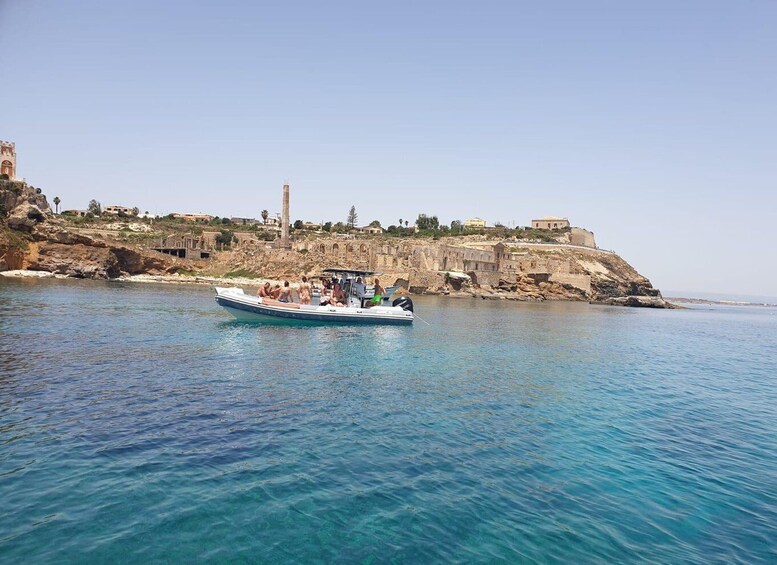 A boat on clear blue water with a rocky coastline and historical buildings in the background. 1