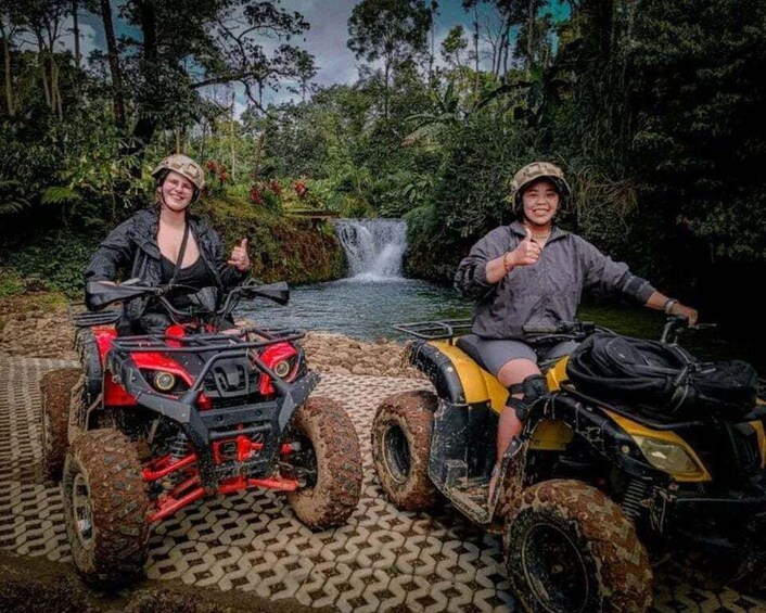 Two people on ATVs in a forested area with a waterfall in the background. 1