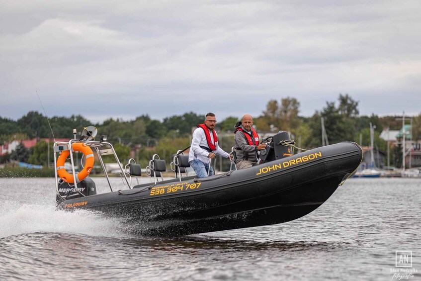 A black inflatable boat named JOHN DRAGON moving on water with two people on board. 1