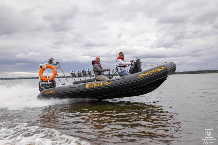 A black speedboat with two people, one driving and one seated, on a body of water with a cloudy sky in the background. 4