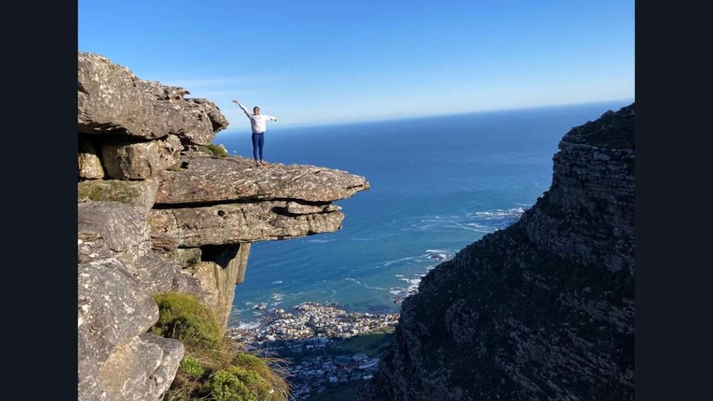 A person standing on a cliff edge with arms outstretched, overlooking a coastal town and the ocean. 1