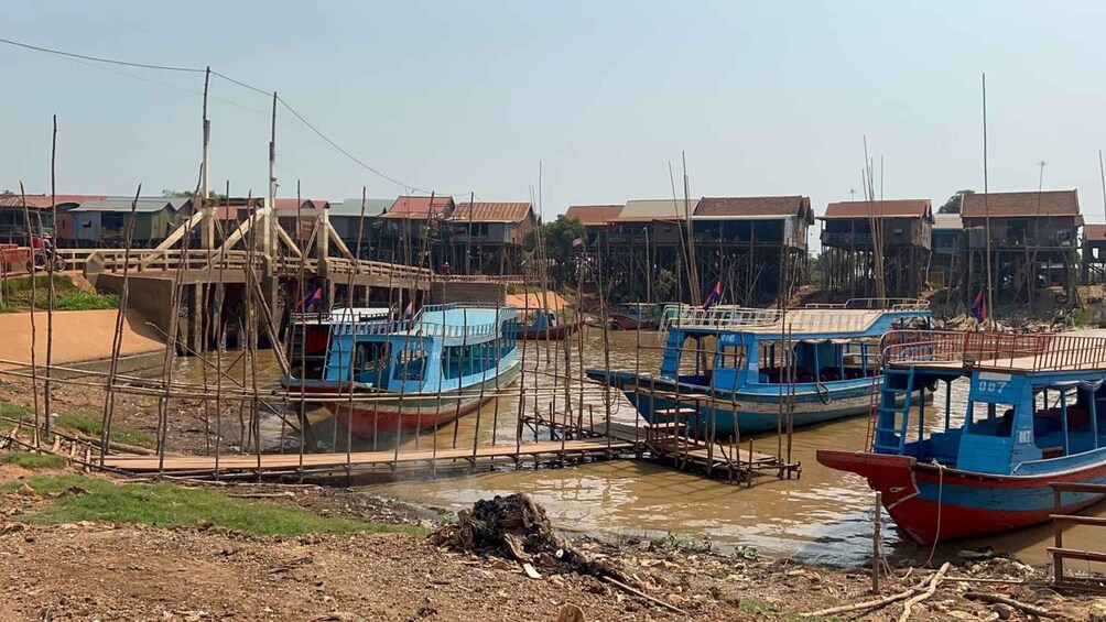 A riverside with several boats docked, a wooden bridge, and residential buildings in the background. 2