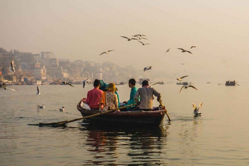 A group of people in a boat on a calm river with birds flying overhead and a cityscape in the background. 1