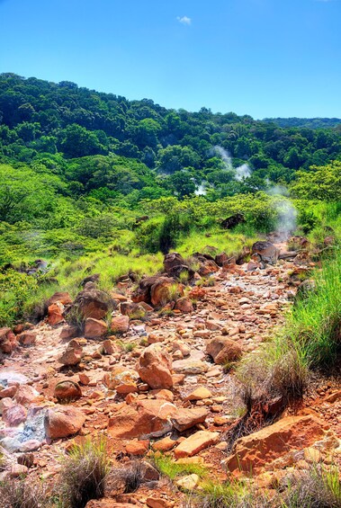 A rocky stream bed with steam rising from the water, surrounded by lush green vegetation and hills in the background. 2