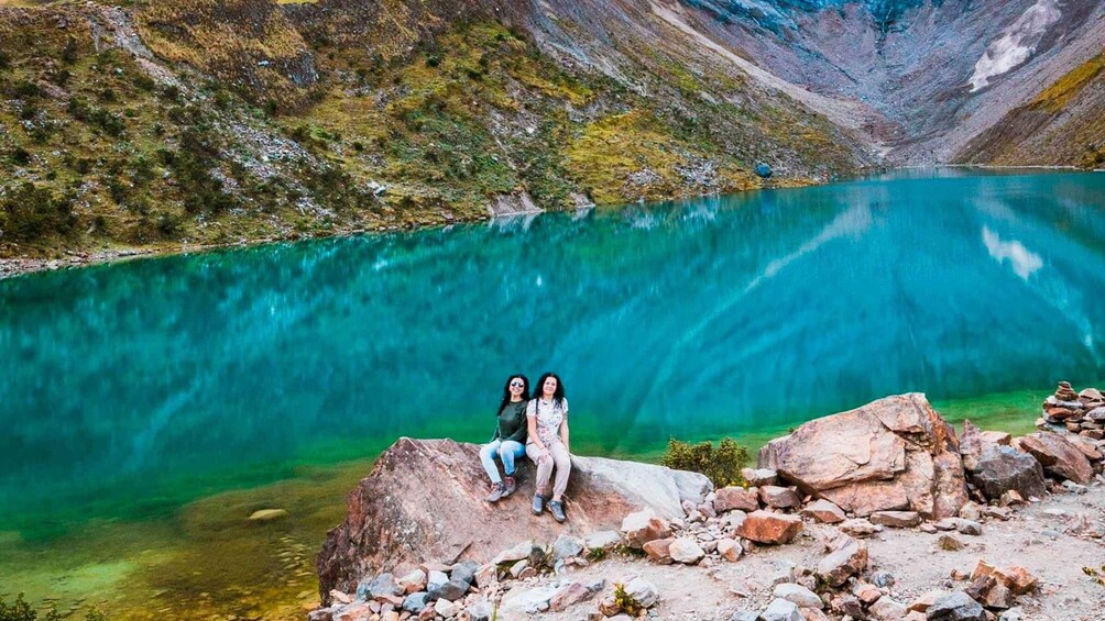 Due persone sedute su una roccia, con un lago turchese e montagne sullo sfondo. 1