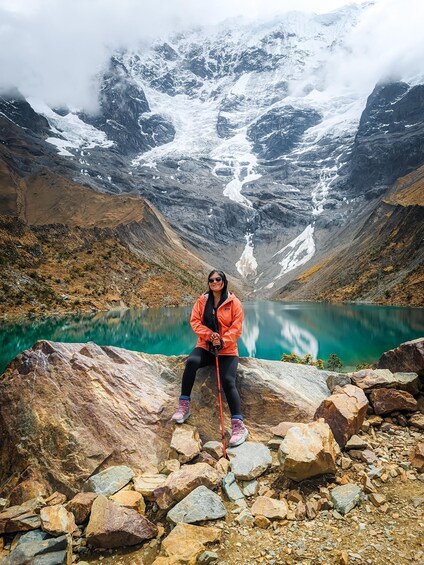 Una persona seduta su delle rocce in riva a un lago turchese, con montagne innevate sullo sfondo. 4