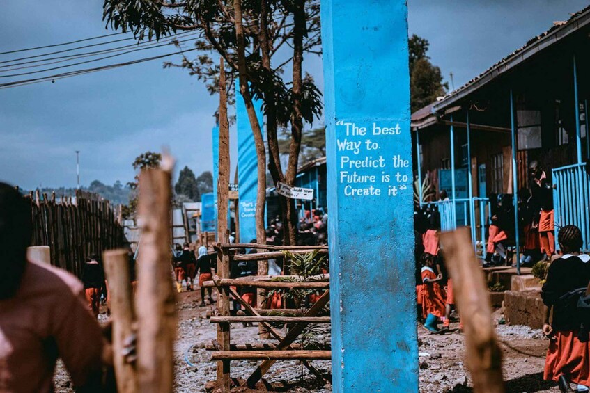 A blue pillar with a quote, a dirt ground, wooden fences, and people in the background. 5