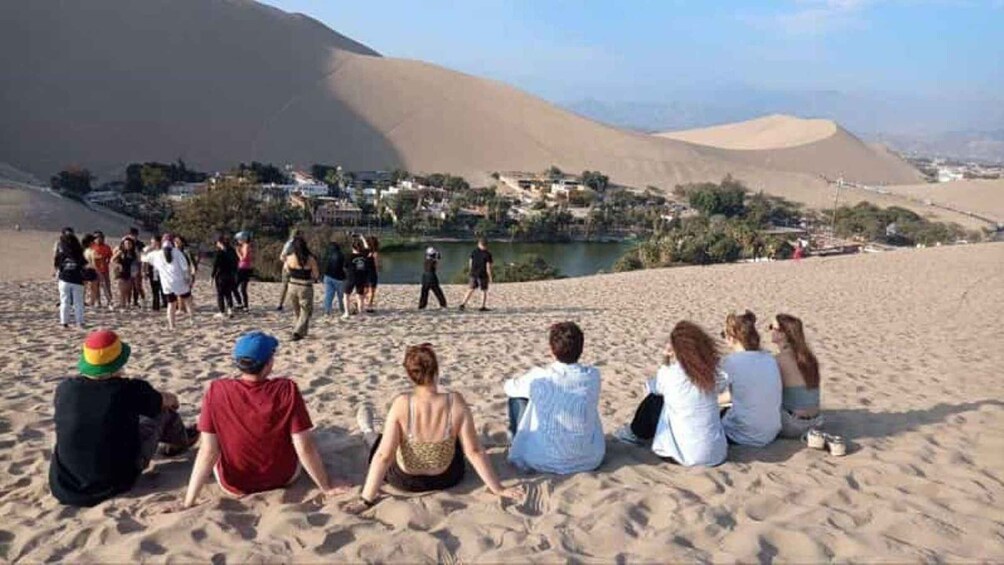 Un grupo de personas sentadas en una playa arenosa con un pueblo al fondo. 4