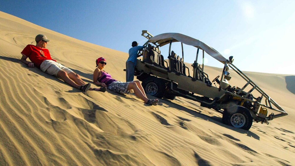 Tres personas descansando sobre una duna de arena, con un buggy aparcado cerca. 3