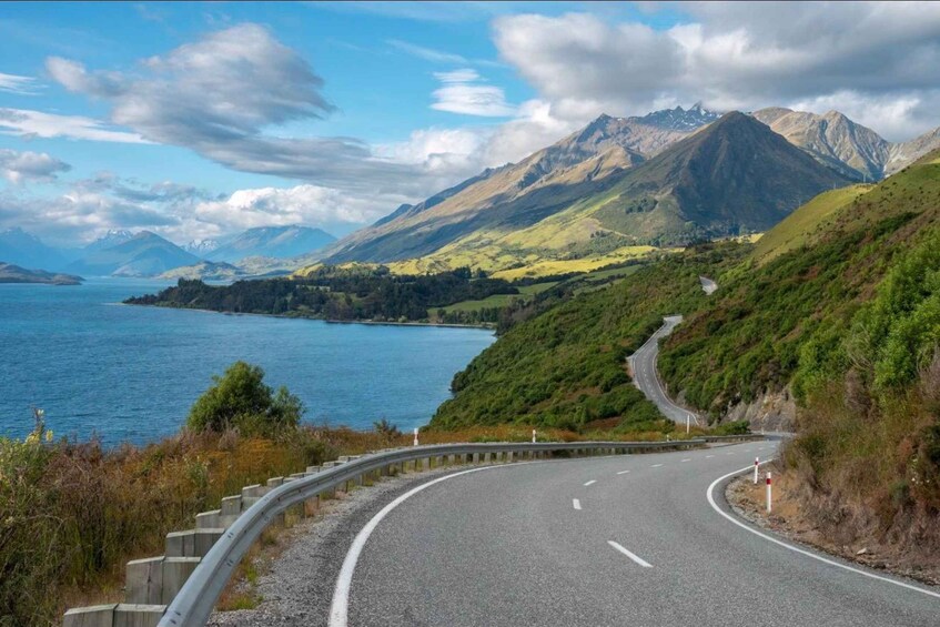 A curving road with a guardrail, surrounded by green hills and a body of water with mountains in the background. 2