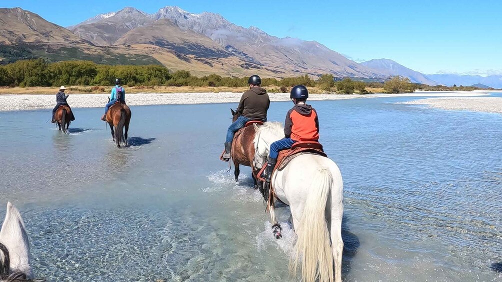 People horseback riding across a partially frozen river with mountains in the background. 5