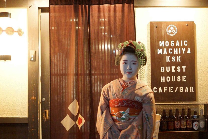 A person in traditional Japanese attire standing in front of a sign that reads "Mosaic Machiya KSK Guest House Cafe/Bar". 5