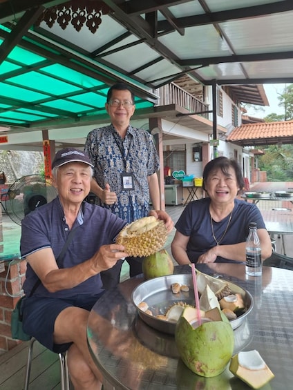 Three people at an outdoor dining area with a table, a fan, and coconuts. 4