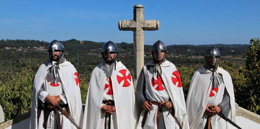 Four people in medieval knight costumes with swords, standing in front of a large cross. 5
