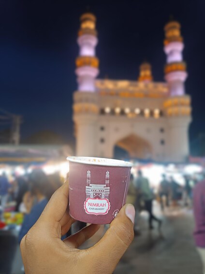 A hand holding a cup with a mosque illustration and the text "NIMRAH CAFE & BAKERY" against a blurred background of a large, illuminated building with two minarets. 5