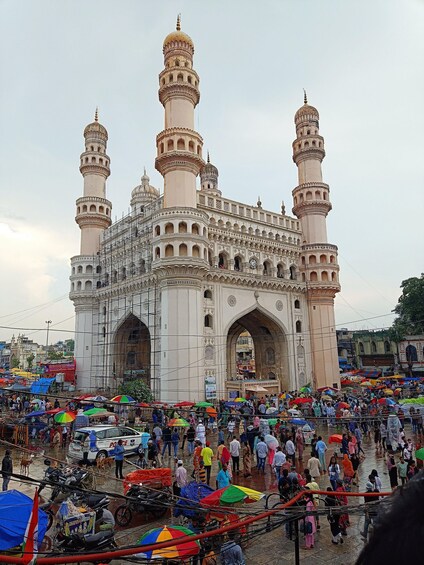 A historic building with multiple minarets and arches, surrounded by a bustling market with people and colorful umbrellas. 3