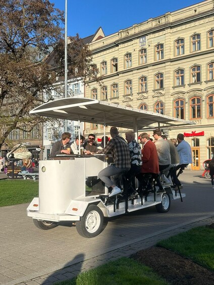 A group of people seated on a white trailer-like structure with a canopy, parked on a paved area in front of a building with ornate architecture. 2