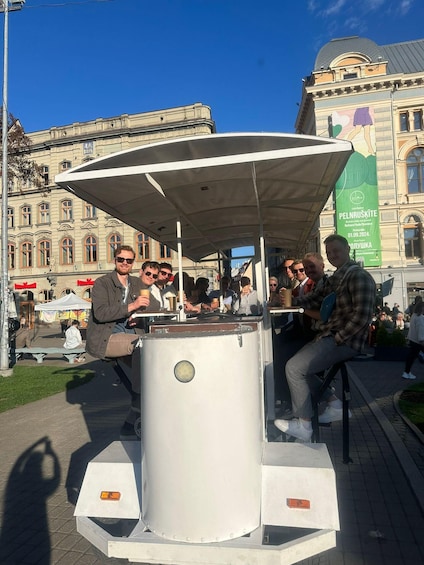 A public square with a kiosk, people sitting and standing, and a large building with classical architecture in the background. 3