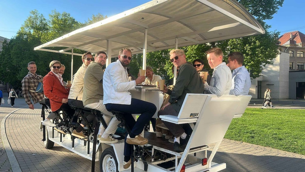 A group of people sitting on a stationary bike-like structure with a canopy, outdoors in a park. 5