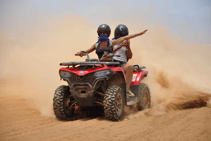 Two people on a red ATV in a sandy desert. 1