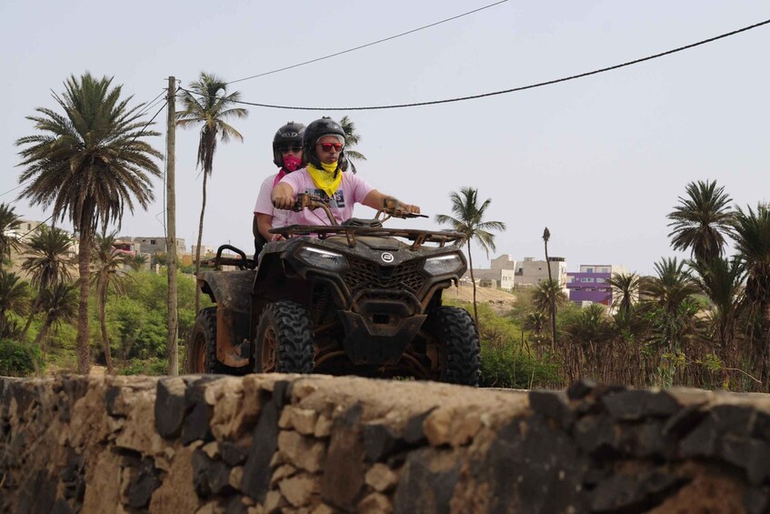 Two people on an ATV in a rural area with palm trees and buildings in the background. 2