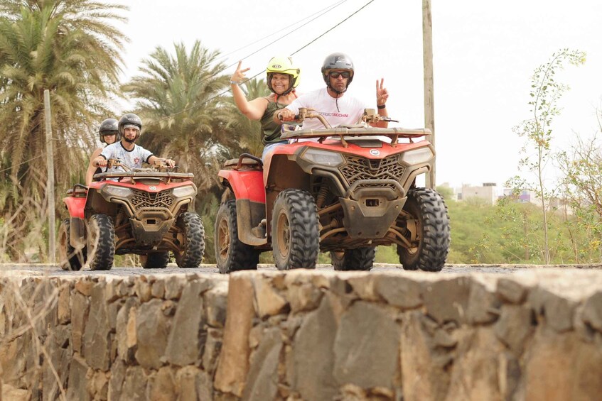 Three people on ATVs on a stone wall with palm trees in the background. 4