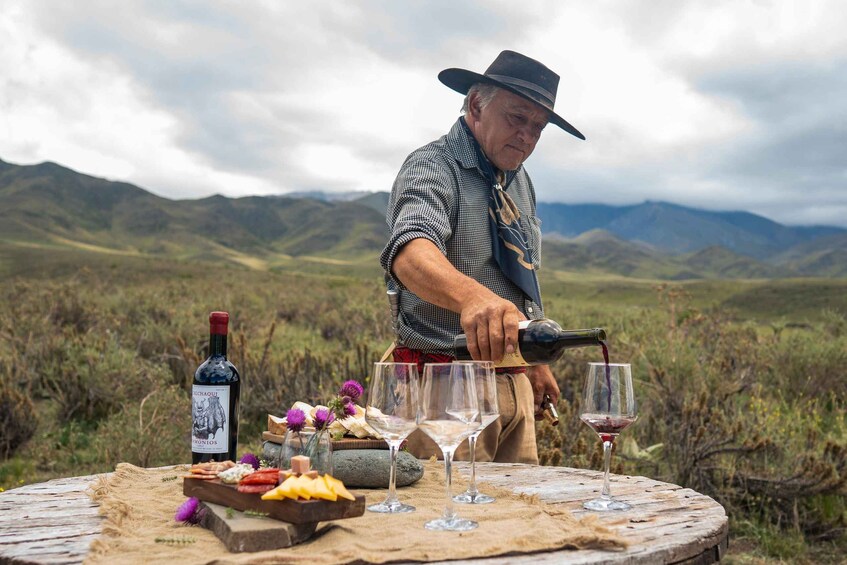 A man pouring wine into glasses at a table set outdoors with a mountainous landscape in the background. 5