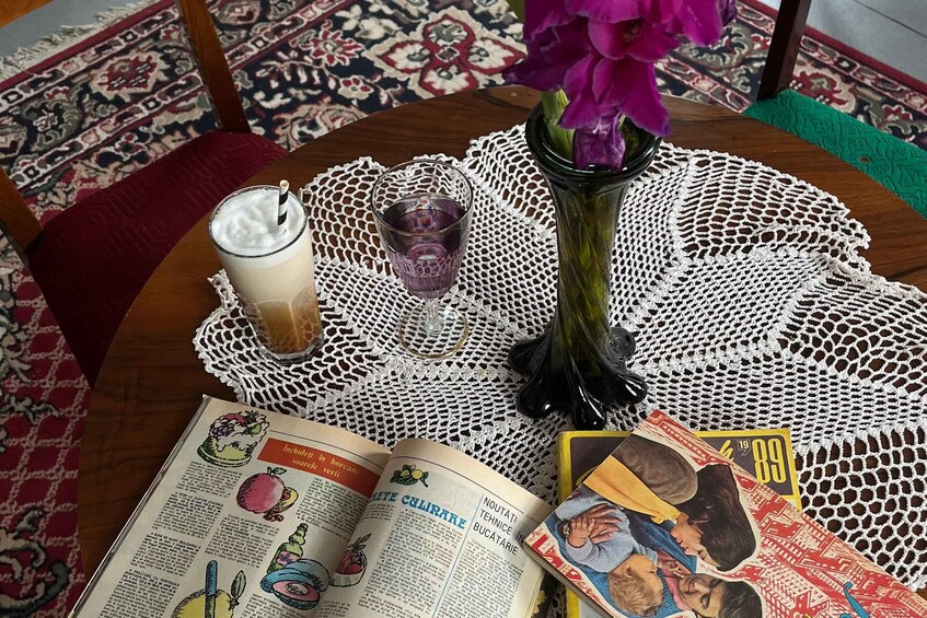 A table with a book, a glass of iced coffee, a glass of water, a vase with purple flowers, and a magazine. 4
