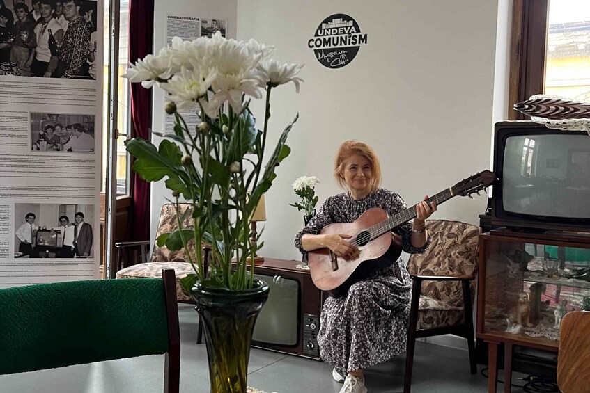 A woman playing a guitar in a room with a vintage television, a vase with flowers, and a wall with a sign. 1