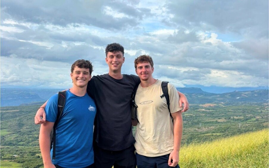 Three people standing on a grassy hill with a mountainous landscape in the background. 2