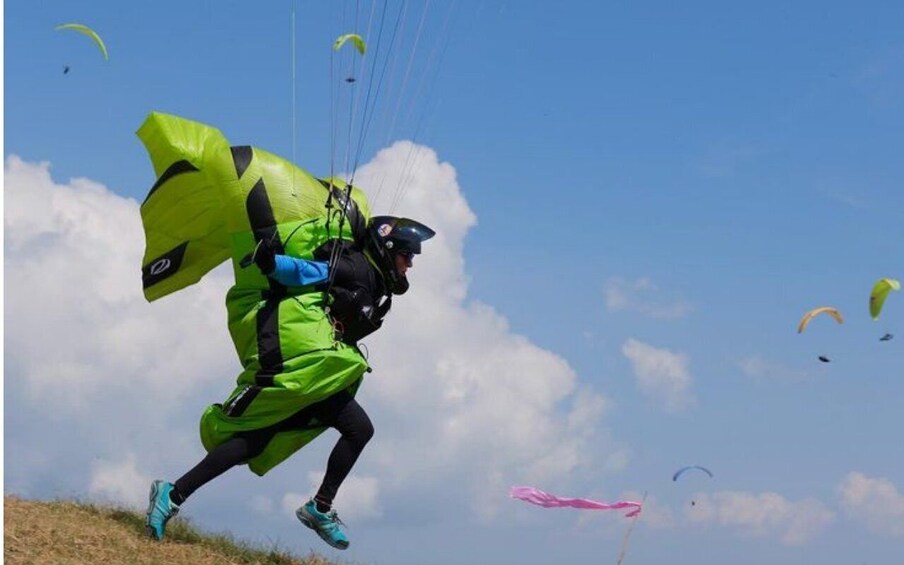 A person paragliding with a bright green parachute, wearing a helmet and goggles, against a clear blue sky with clouds. 3