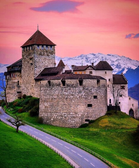 A historic stone castle with a tower, surrounded by lush green hills and a mountain range in the background. 1